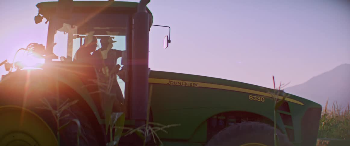 Two people ride a green tractor through a field at sunrise