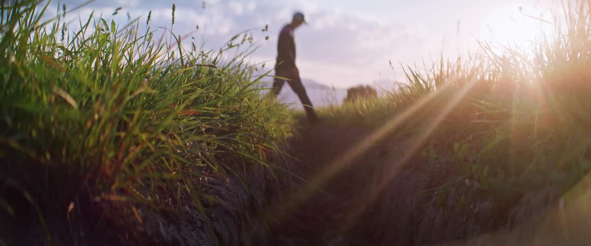Man silhouetted by the glaring sun steps over a ditch in a field on a cloudy day