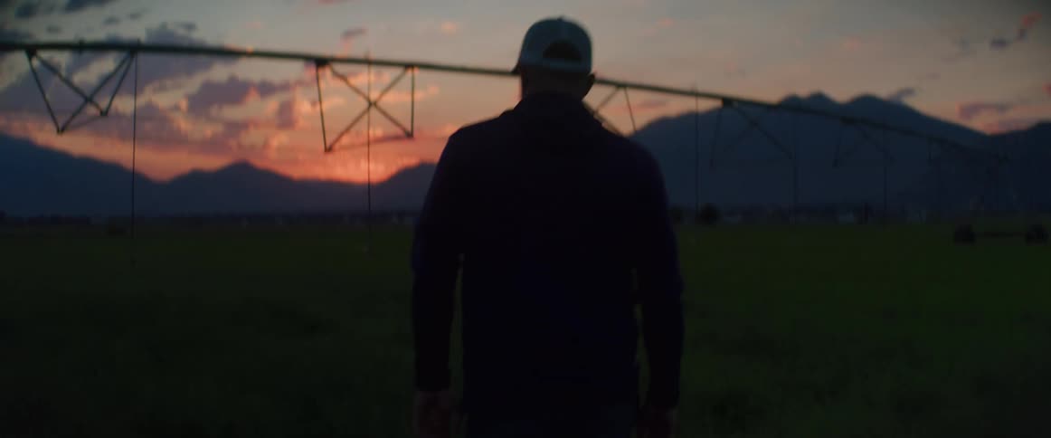 A man walks through a green field at sunset with mountains in the background