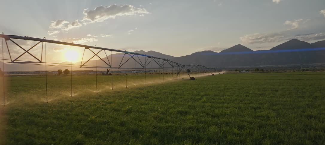 A centerpivot irrigation system operates spraying streams of water onto a large field of green growth under a bright sun all against a background of high mountains under a cloudy sky