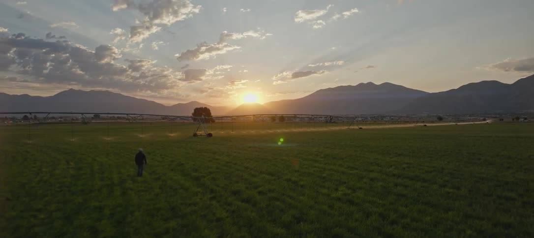 A person walks across a grassy field at sunset with mountains behind