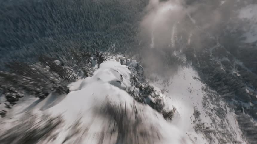 A breathtaking aerial view swoops dramatically over a snow covered mountain range revealing rugged peaks pine forests and clouds under a blue sky