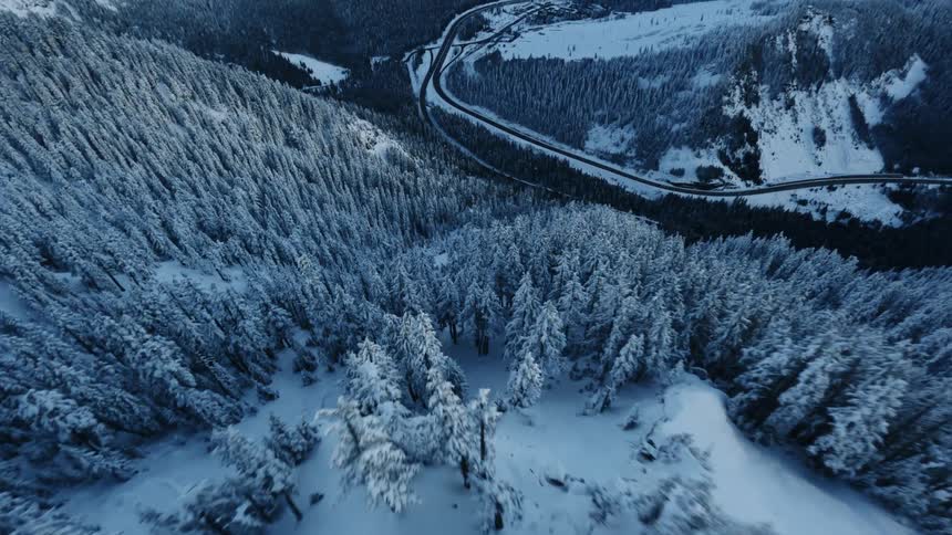 An exhilarating aerial shot descends and reveals a winding highway cutting through snow covered mountain forests under a clear blue sky