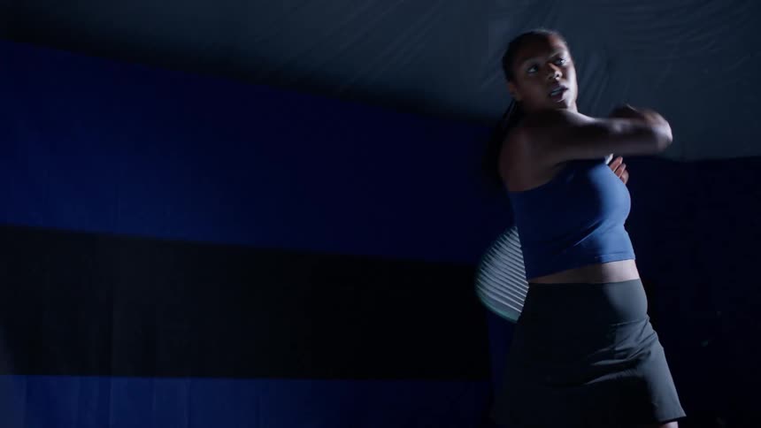 A focused young female tennis player wearing a blue top and black skirt practices her serve on a dimly lit court