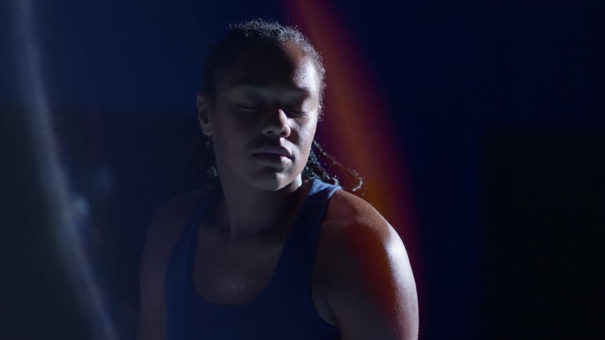 Athletic woman in dim blue light closes eyes sweat glistening expression calm and determined serves a tennis ball using a tennis racket