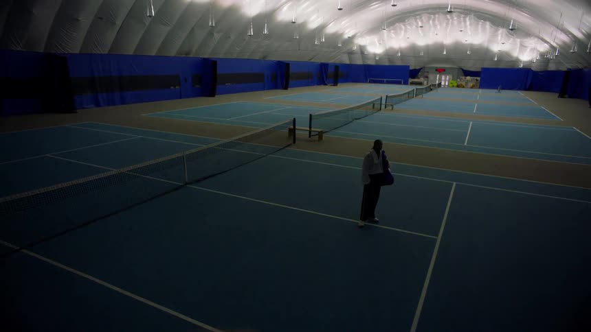A lone tennis player stands on a blue court inside a cavernous softly lit dome