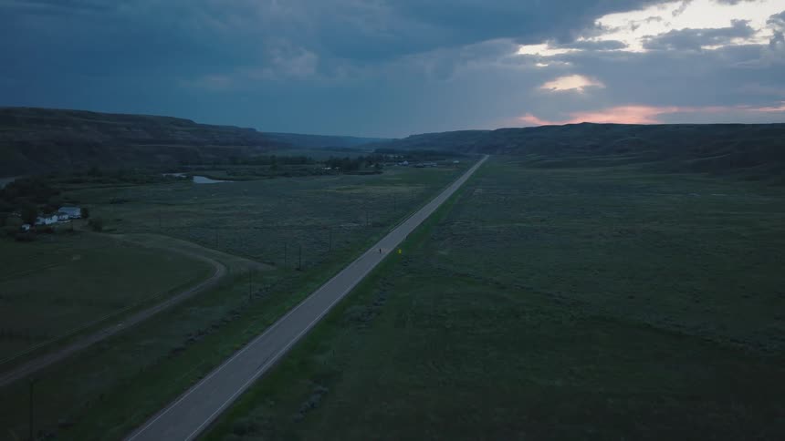 An aerial view shows a person riding a motorcycle along a lonely countryside road at sunset
