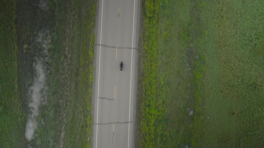 Aerial view view of a motorcyclist riding a narrow rural highway bordered by lush green fields