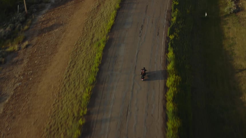 A motorcyclist rides down a cracked rural road between sunlit grassy fields