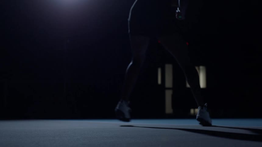 The lower body silhouette of a person in athletic wear and white sneakers performs jumping rope exercises in a dark indoor training space with a red timer display visible in the background