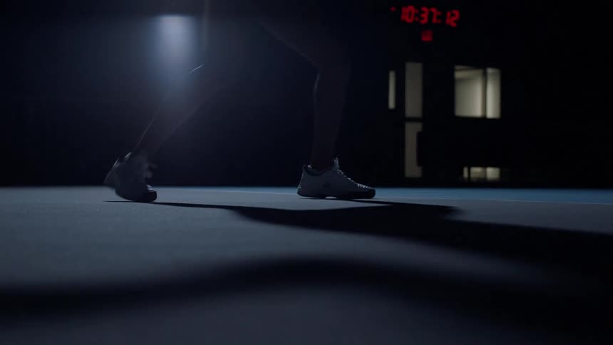 The lower body silhouette of a person in athletic wear and white sneakers performs jumping rope exercises in a dark indoor training space with a red timer display visible in the background