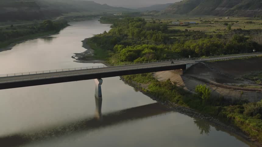 Aerial shot of a lone motorcyclist crossing a low bridge over a calm reflective river at dusk