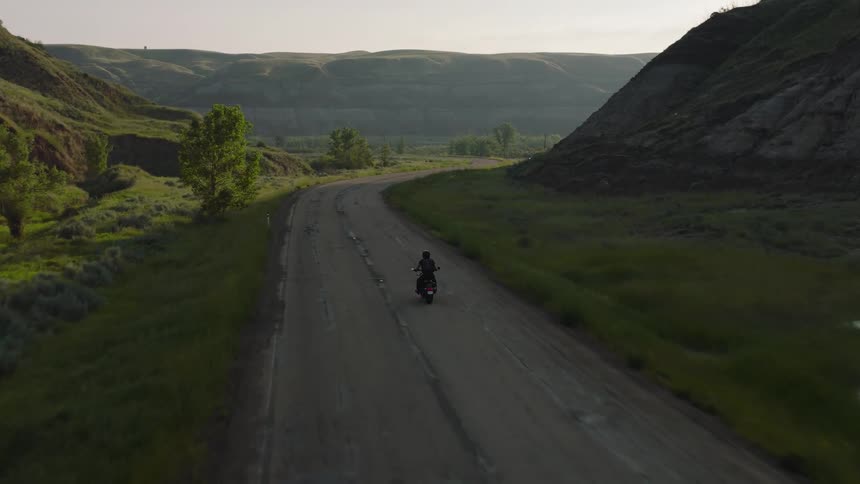 Solitary rider cruises a patched rural road between green valley hills under soft sunlight