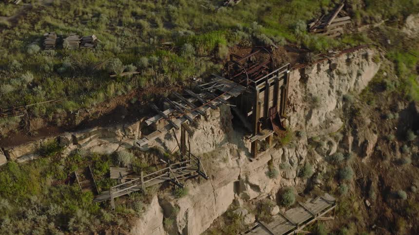An aerial view shows the dilapidated wooden remains of an old mining structure clinging to a steep eroded cliff face covered in sparse green and brown vegetation under daylight