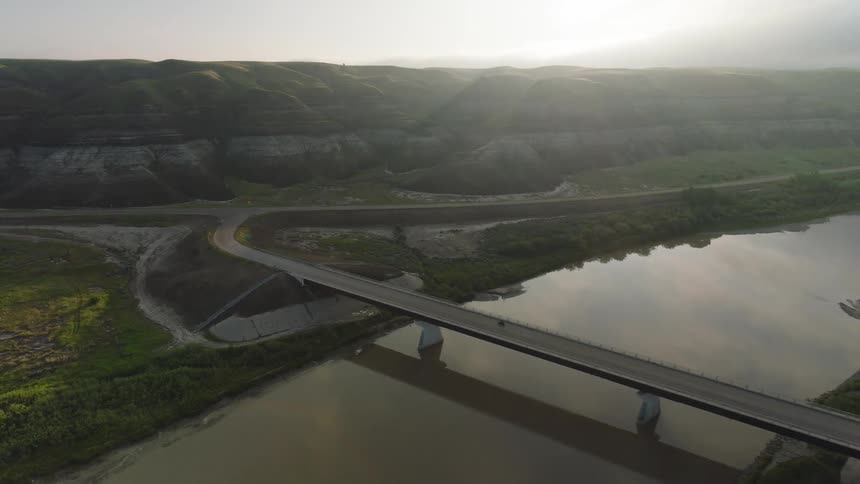 A motorcycle travels across a wide bridge spanning a reflective river beneath hazy sunrise light hills layered beyond