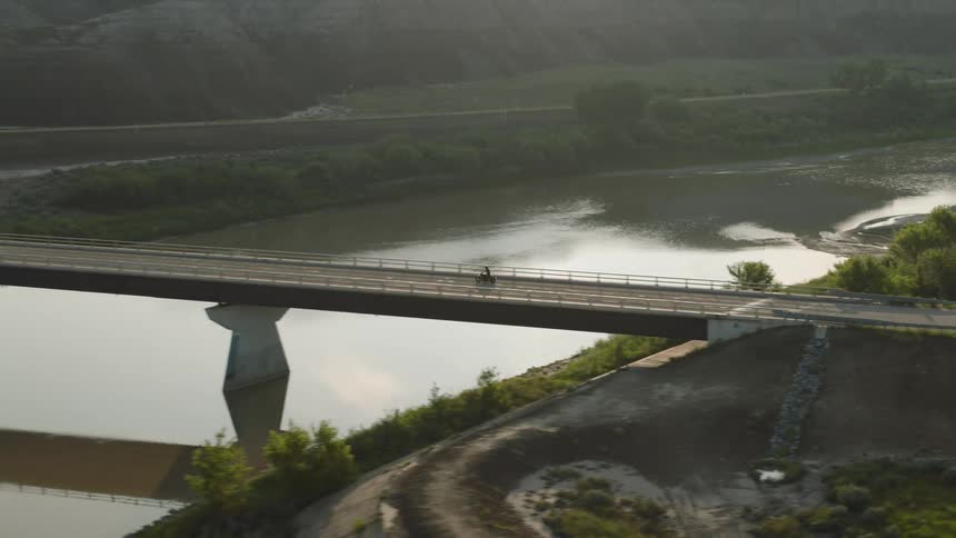 A person rides a motorcycle across the countryside crossing a bridge at sunrise