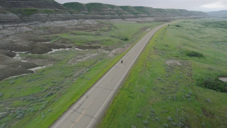 A lone motorcycle traveling down a straight paved road on a green terrain under a cloudy overcast sky
