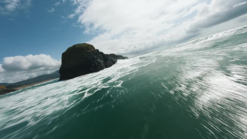 An action camera view captures a large turquoise ocean wave breaking with white foam against a dark coastal sea stack with green forested cliffs and a partly cloudy blue sky in the distance