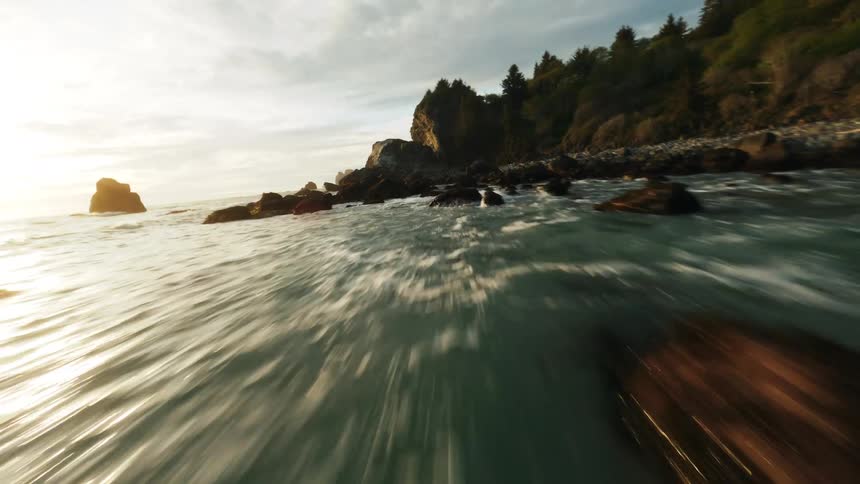 Low waves glide toward a rocky coastline under soft sunset light with forested cliffs rising ahead