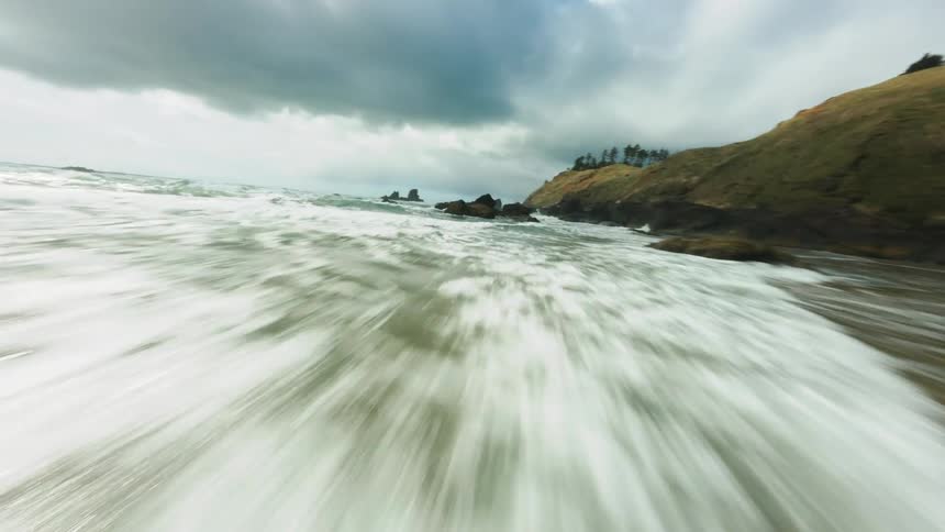 A dynamic lowangle shot rapidly moves across a wet brown sandy beach as a foaming ocean wave rushes in with rocky cliffs and pine trees visible under a dramatic cloudy sky