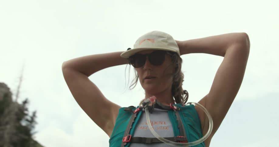 Woman wearing a cap sunglasses and a hydration backpack looks at the landscape with hands on their head