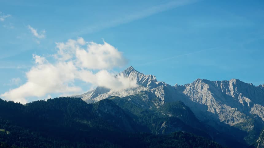 A rugged tall grey mountain range with visible jagged peaks partially covered by moving white clouds rising above a dark green pinecovered base against a backdrop of a bright clear blue sky