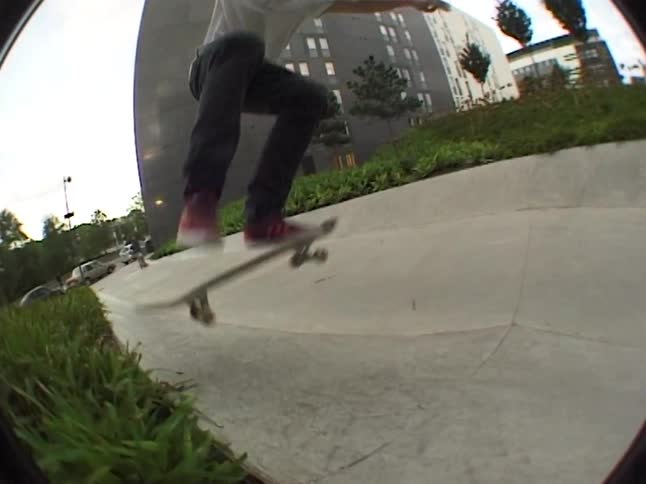 A young man wearing a white polo shirt and dark pants performs a trick on a skateboard in a concrete urban park with a large building and overpass in the background captured with a fisheye lens