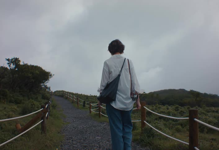 A young woman with short dark hair wears a white shirt and blue jeans while walking away along a gravel path lined with rope fences on a misty hillside on an overcast day