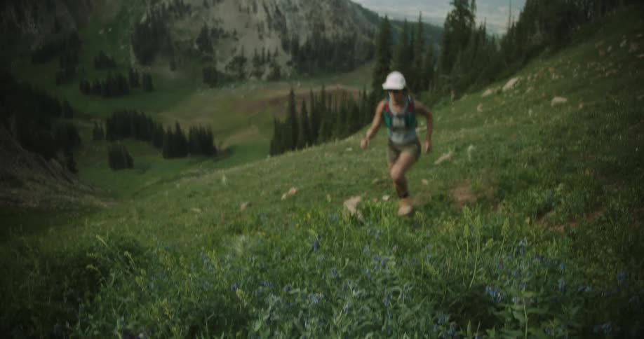 A woman trail runner climbs a steep alpine meadow looking focused and determined beneath jagged mountain peaks