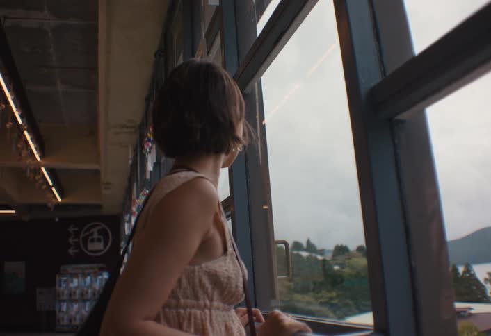 A woman approaches the wall of a building made entirely of glass sections including doors and windows and looks to the view of a river houses and a mountain