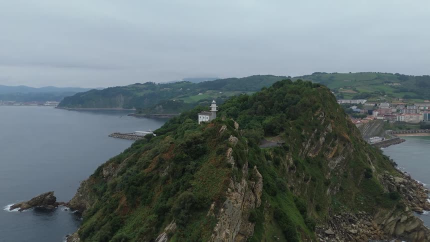 An old lighthouse with a house next to it on top of a mountain peninsula surrounded by forest trees