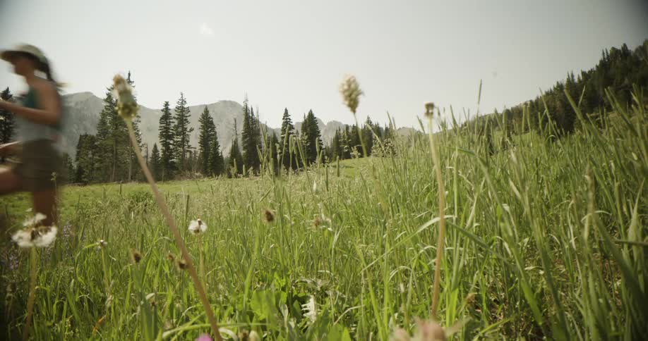 A woman runs joyfully downhill through a sunlit alpine meadow of tall grass and wildflowers with mountains beyond