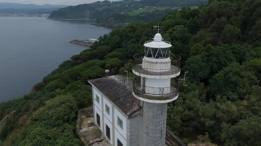 An old lighthouse with a house next to it on top of a mountain peninsula surrounded by forest trees