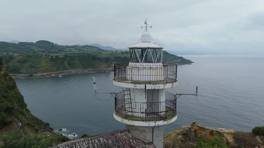 A weathered white lighthouse rises above a rugged coastline overlooking the calm sea and rolling green hills
