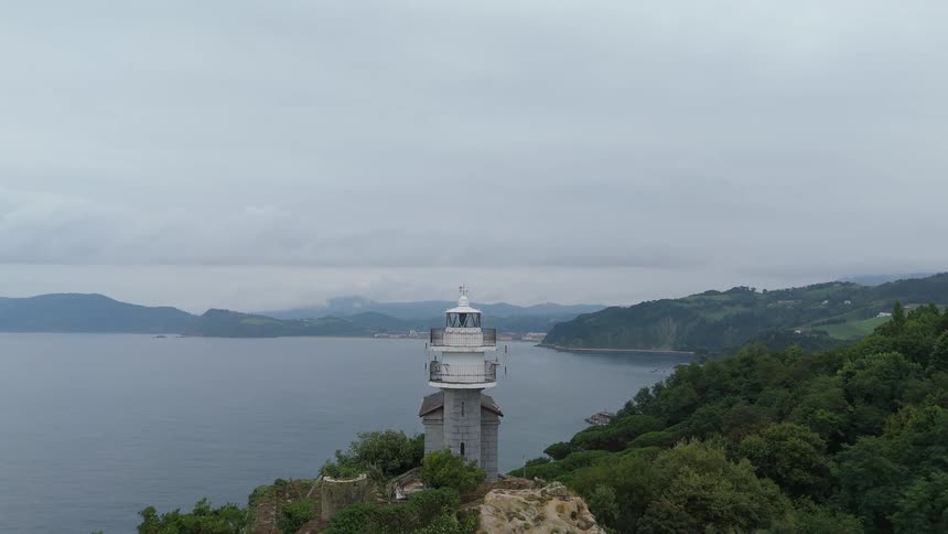 A solitary lighthouse crowns a rocky ridge above the bay framed by forested hills under a gray sky
