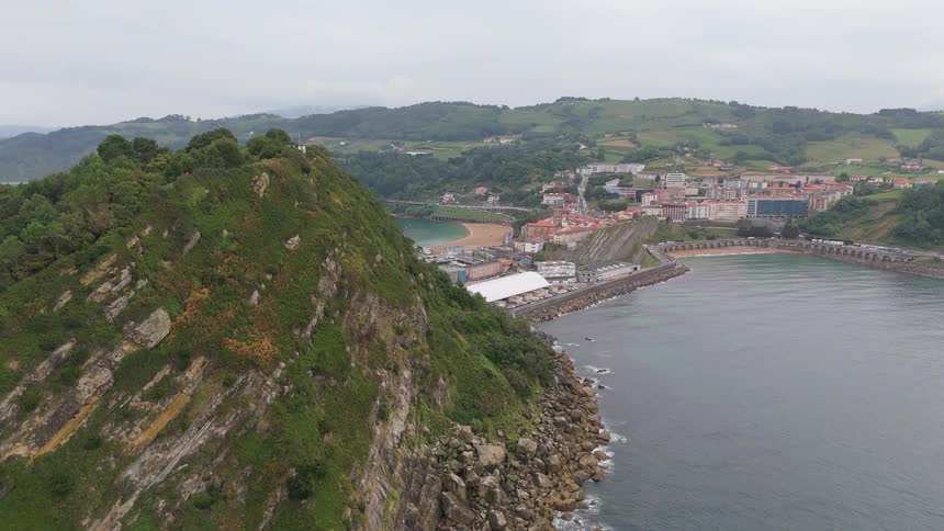 Cliff covered with vegetation beside a coastal town with a marina and beach in the background