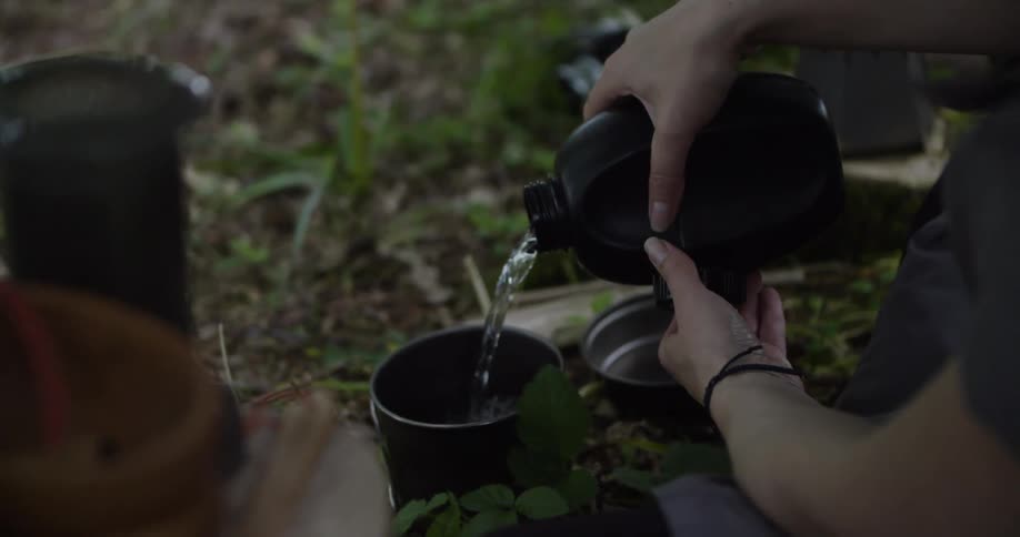 Person pours water from a canteen into a metal container on a leafcovered ground