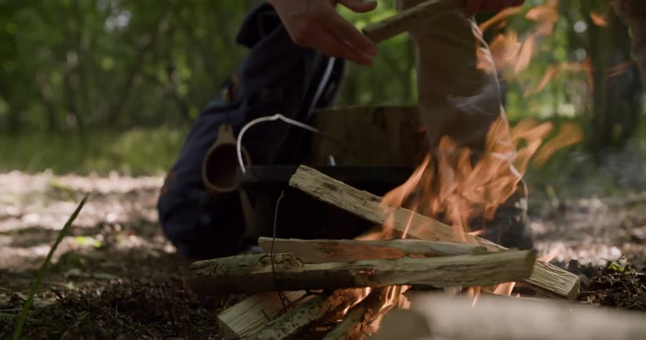 A young man builds a campfire with wood while camping in the forest at midday surrounded by trees