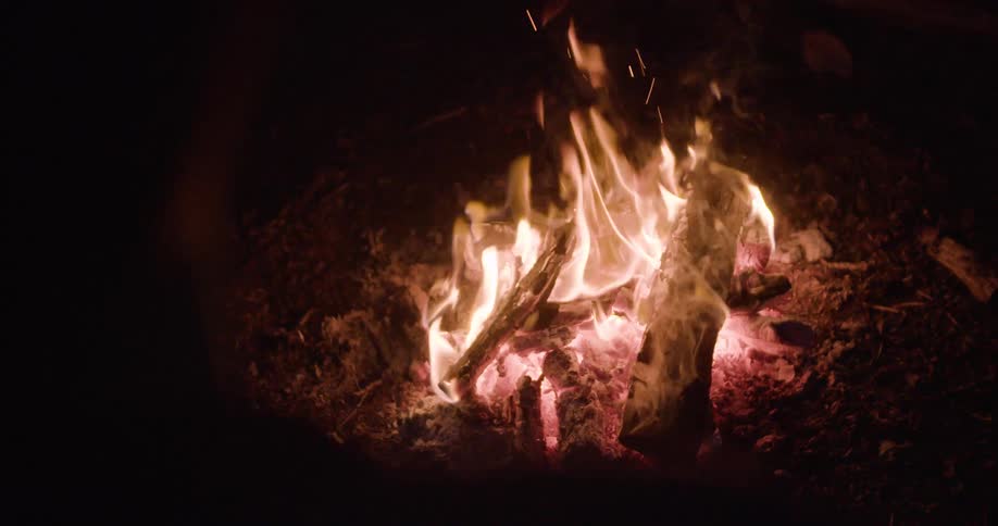 Orange and yellow flames engulf several pieces of wood resting on glowing red embers and dark earth with small sparks ascending from the heat source in an otherwise dark environment