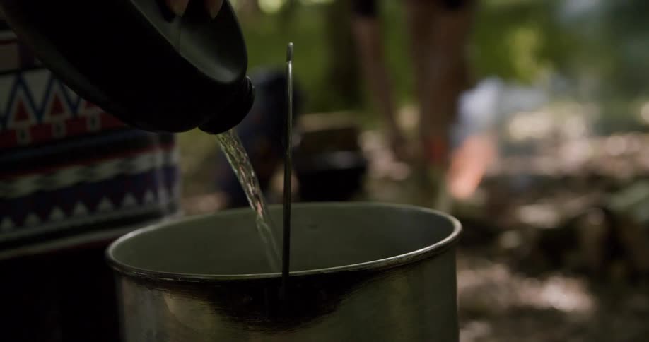 A person pours a stream of clear liquid from a dark plastic bottle into a large round metal pot with a wire handle while another person is visible in the blurry background tending to a small burning fire on the forest floor