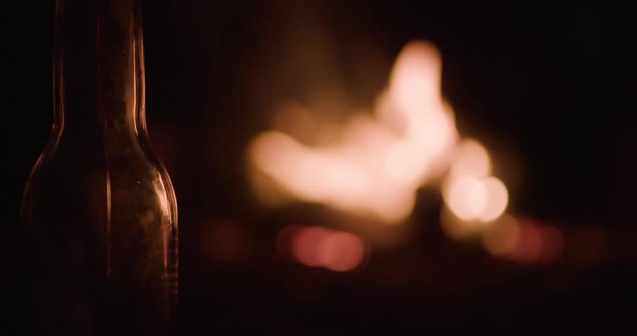 A close shallow depth of field view shows the silhouette of a glass bottle in the foreground with a blurry bright orange campfire burning in the dark background