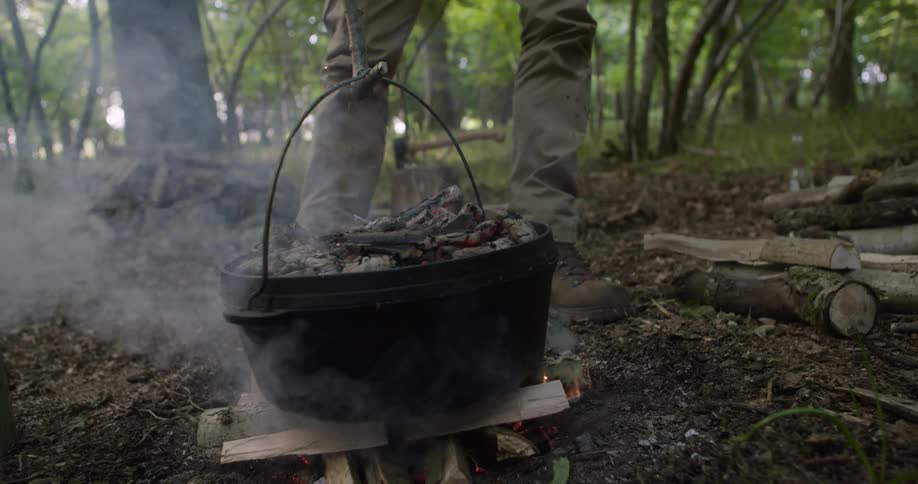 The man is placing a large pot of food on the burning logs