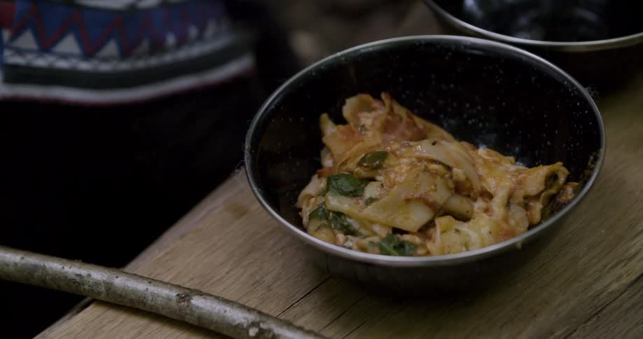 Bowl with a serving of pasta and vegetables rests on a wooden surface outdoors