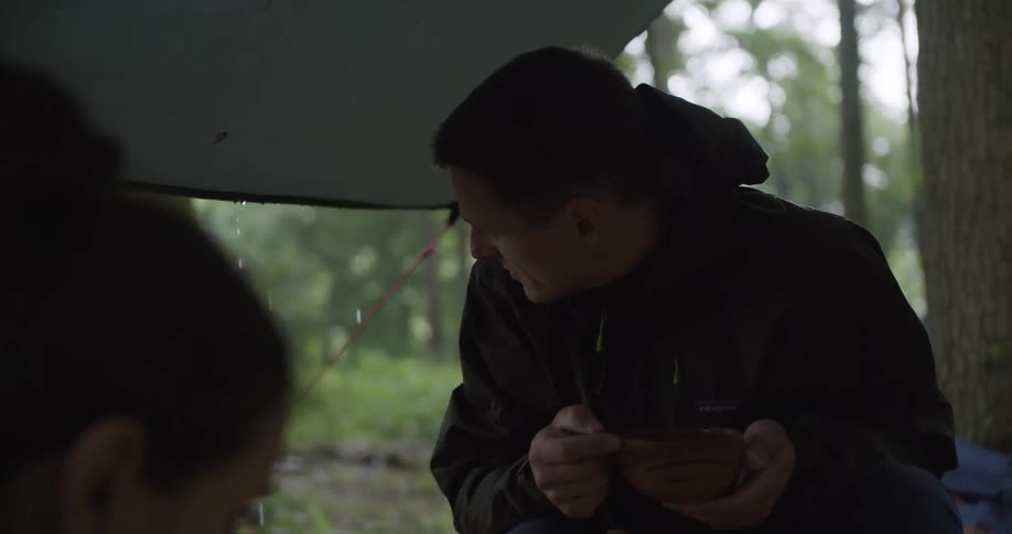 A man wearing a black jacket holds a wooden bowl in both hands while sitting under a dark tarp with drops of water visible with another persons outoffocus head in the immediate foreground and a treefilled forest visible in the background
