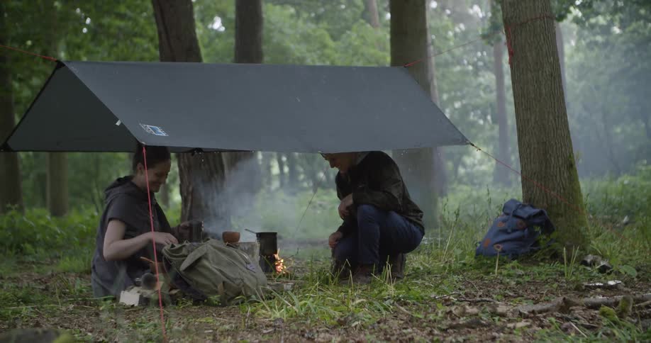 A couple sits under a tent in the woods at midday cooking together while camping as sunlight filters through the trees