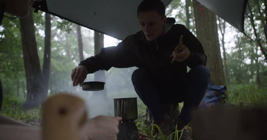 A man wearing a dark jacket squats under a raised tarp in a forest and uses a small dark vessel to pour something from a metal mug sitting on a small stove handing the vessels contents toward a second person in the immediate foreground