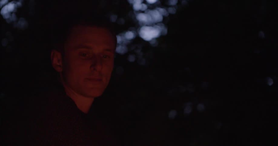 A young man sitting by a campfire outdoors in a woodland area at night