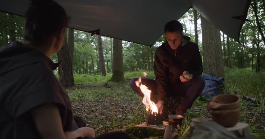 Two people sit on the forest floor under a lowhanging dark tarp with a small metal woodburning stove with a large flame between them as both warm hands with dark forest trees visible in the background