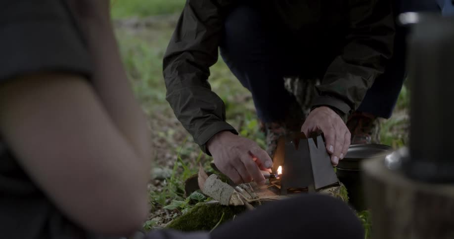 A young man sets up a campfire in the woods while camping as a woman stands to the side watching him