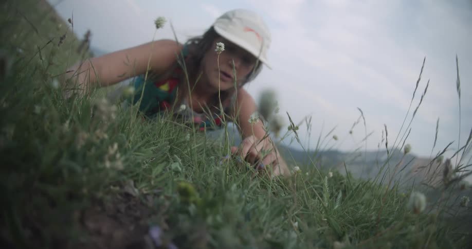 A woman hikes up a grassy hill at midday wearing a hat but suddenly loses her balance and falls to the ground